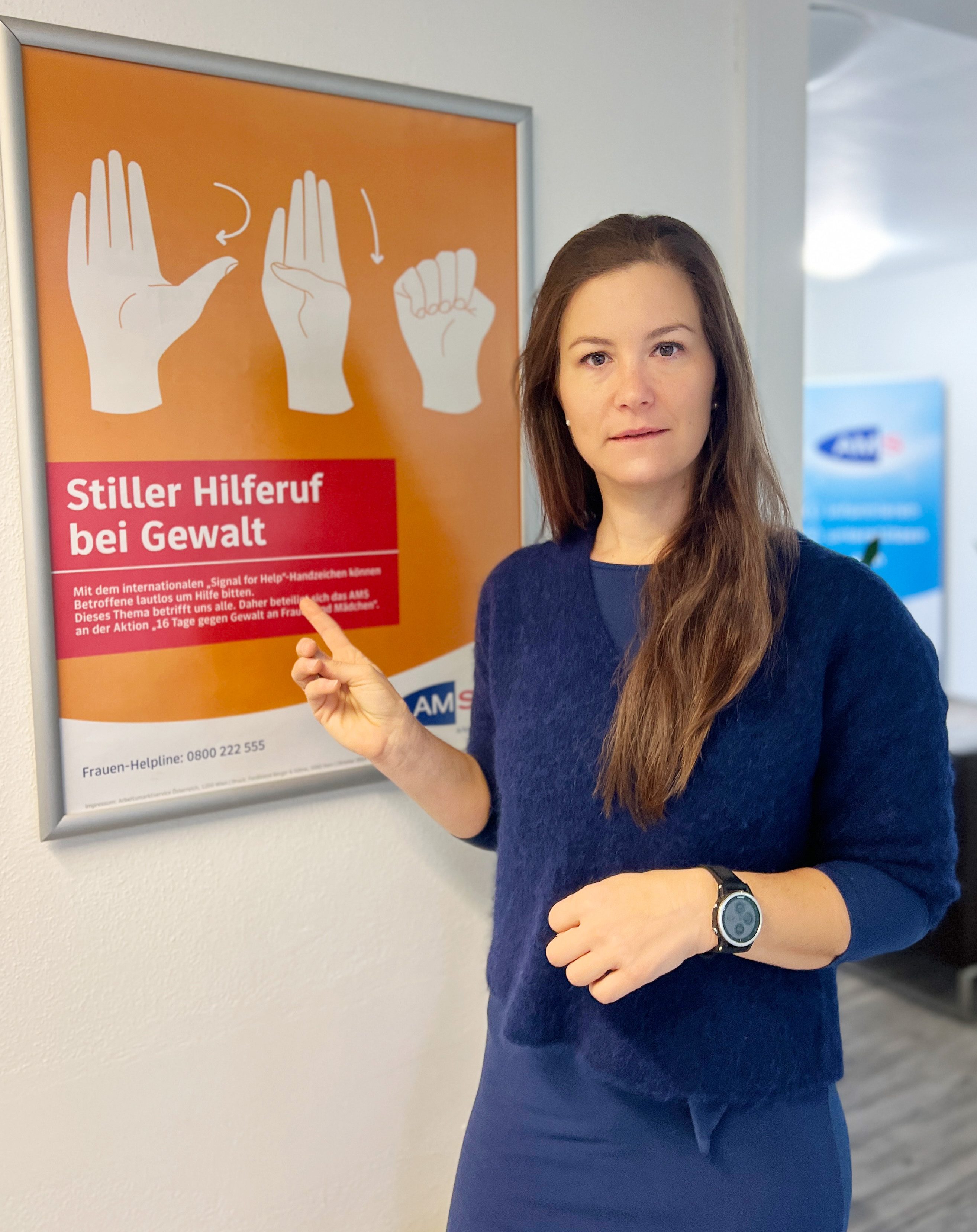Julia Kröll, interimistische Landesgeschäftsführerin des AMS Salzburg, vor dem orangefarbenen Plakat mit dem „stillen Hilferuf bei Gewalt“