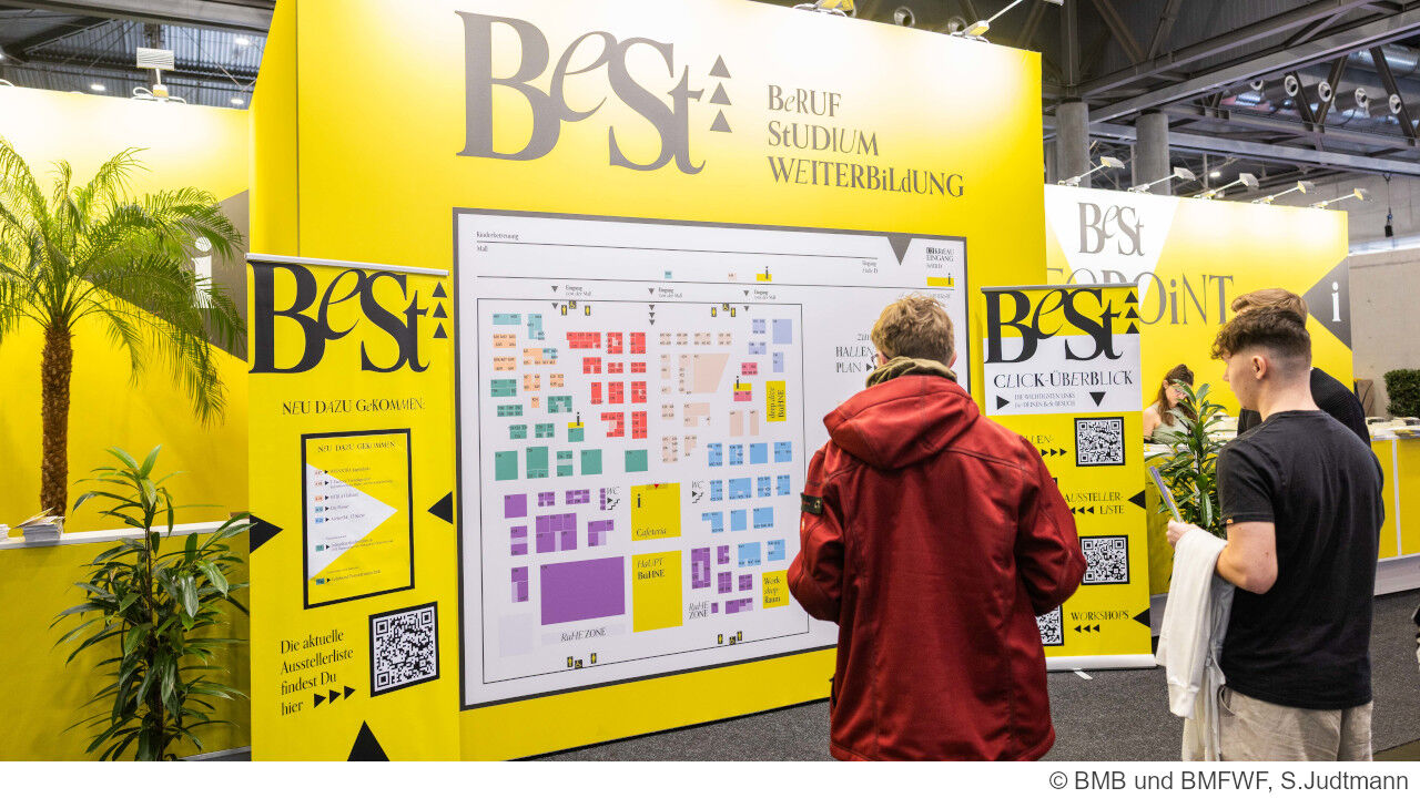 Zeigt den Eingangsbereich der BeSt Messe mit großem gelben Aufsteller mit dem BeSt Logo und Lageplan der Messe. Davor stehen drei Junge Leute die sich den Lageplan ansehen.