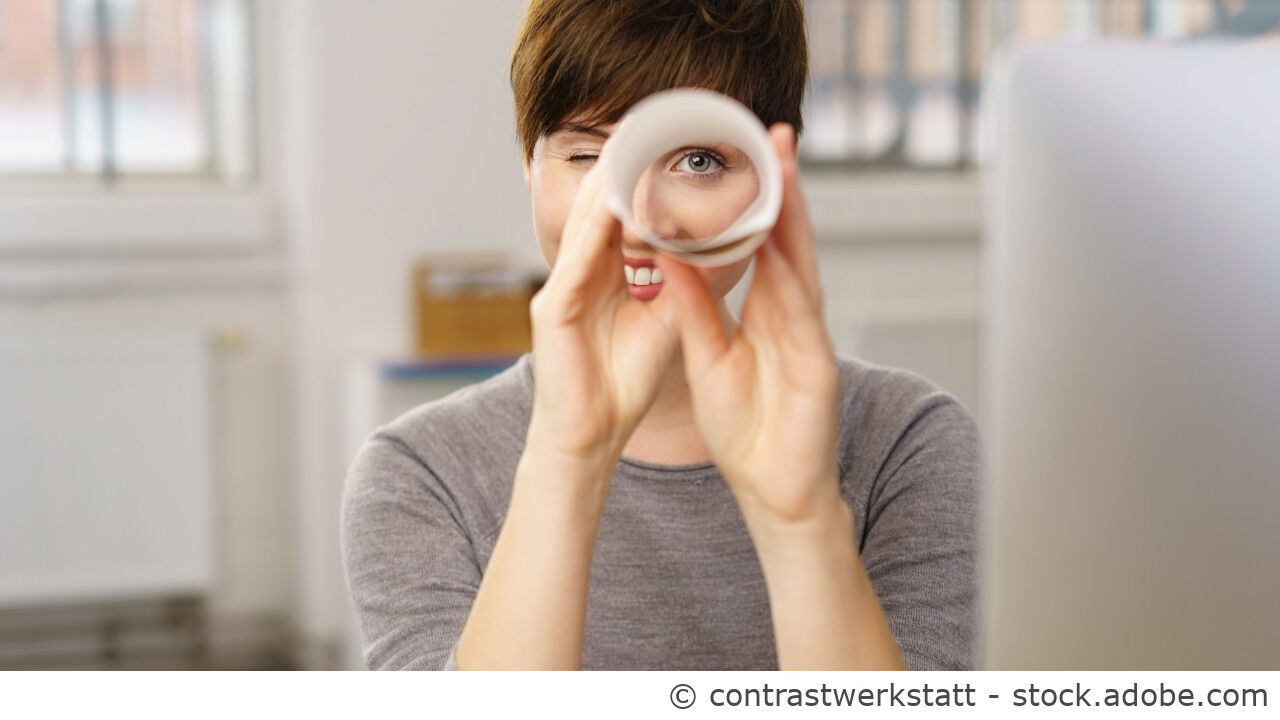 Portrait einer jungen Frau die mit einer Papierrolle als Fernglas in die Kamera schaut.